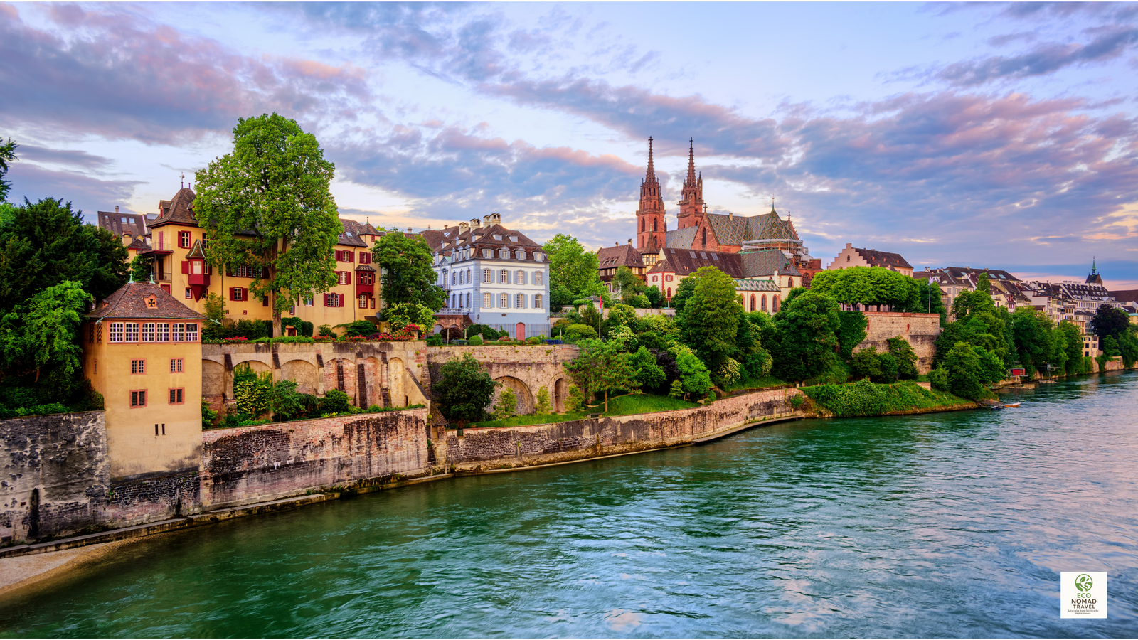 Basel’s Rhine River waterfront with historic old buildings, leafy trees, and reflections on the water in Switzerland.