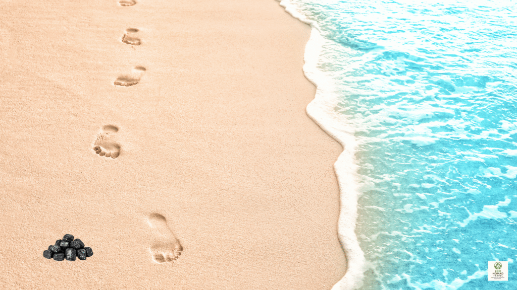 Footprints on a bright sand beach next to a small pile of black carbon chunks, symbolizing a traveler’s carbon footprint on a blue-water coastline