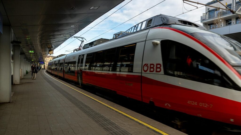 A sleek red and white ÖBB train on a platform at a modern station, with passengers waiting.