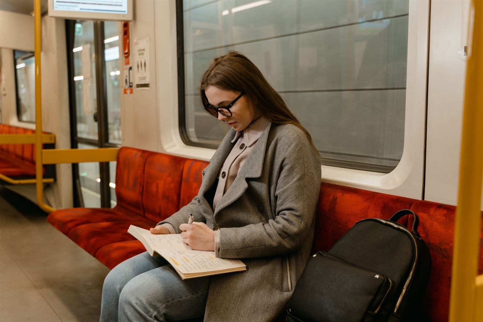 A woman sitting alone on a train, writing in a notebook with a backpack on the seat beside her
