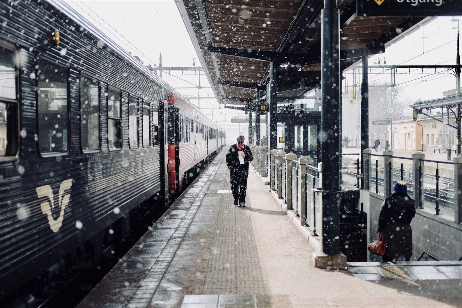 A train waiting at a station platform during heavy snowfall, with passengers walking alongside